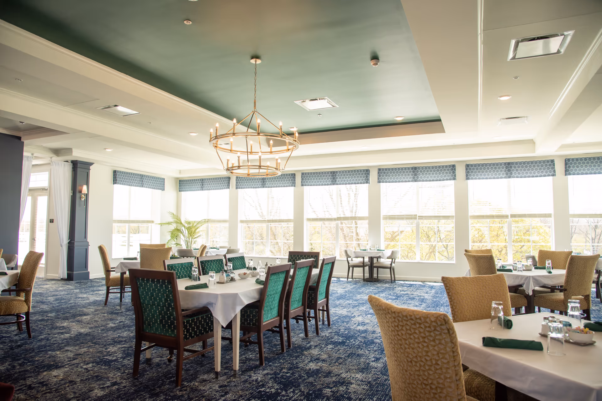 A bright and spacious dining room with large windows letting in natural light. The room features multiple tables covered with white tablecloths, set with glasses, napkins, and condiments. Chairs with green and beige upholstery surround the tables. A large, circular chandelier hangs from the ceiling, which is painted green in the center. The carpet is blue with a patterned design, and there are decorative window valances above the windows.