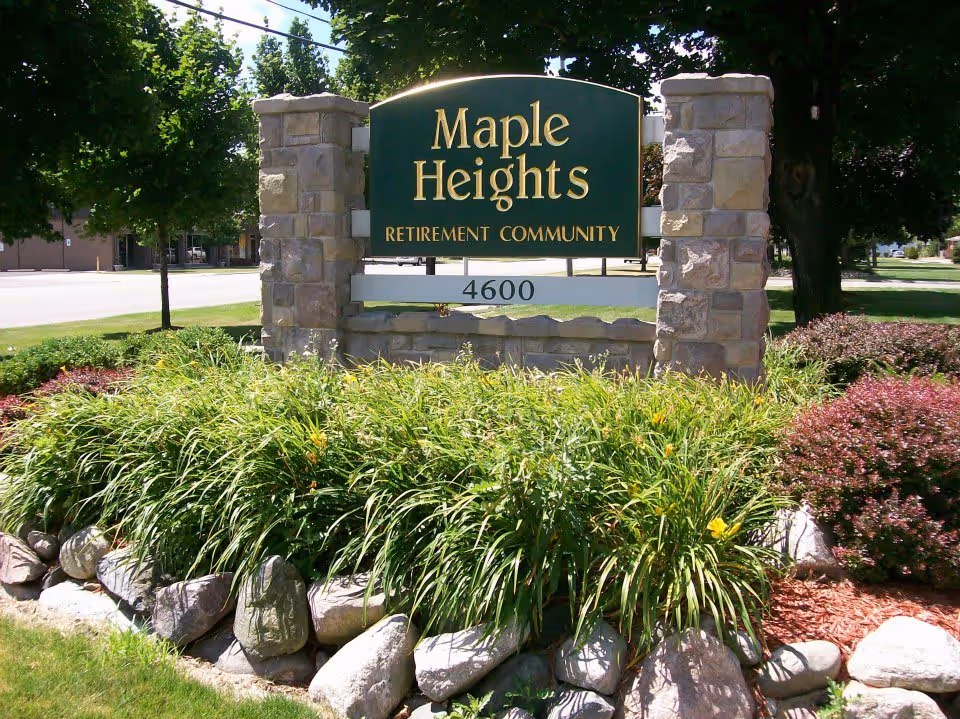Stone sign for Maple Heights Retirement Community with the address number 4600, surrounded by green plants, bushes, rocks, and trees in a landscaped outdoor area.