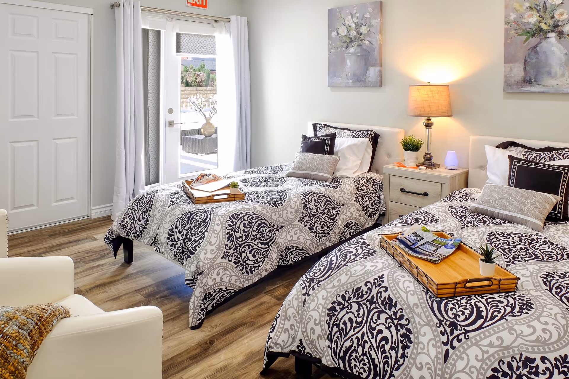 Bright twin bedroom with patterned bedding, a nightstand and lamp, trays on the beds, and a glass door leading outside.