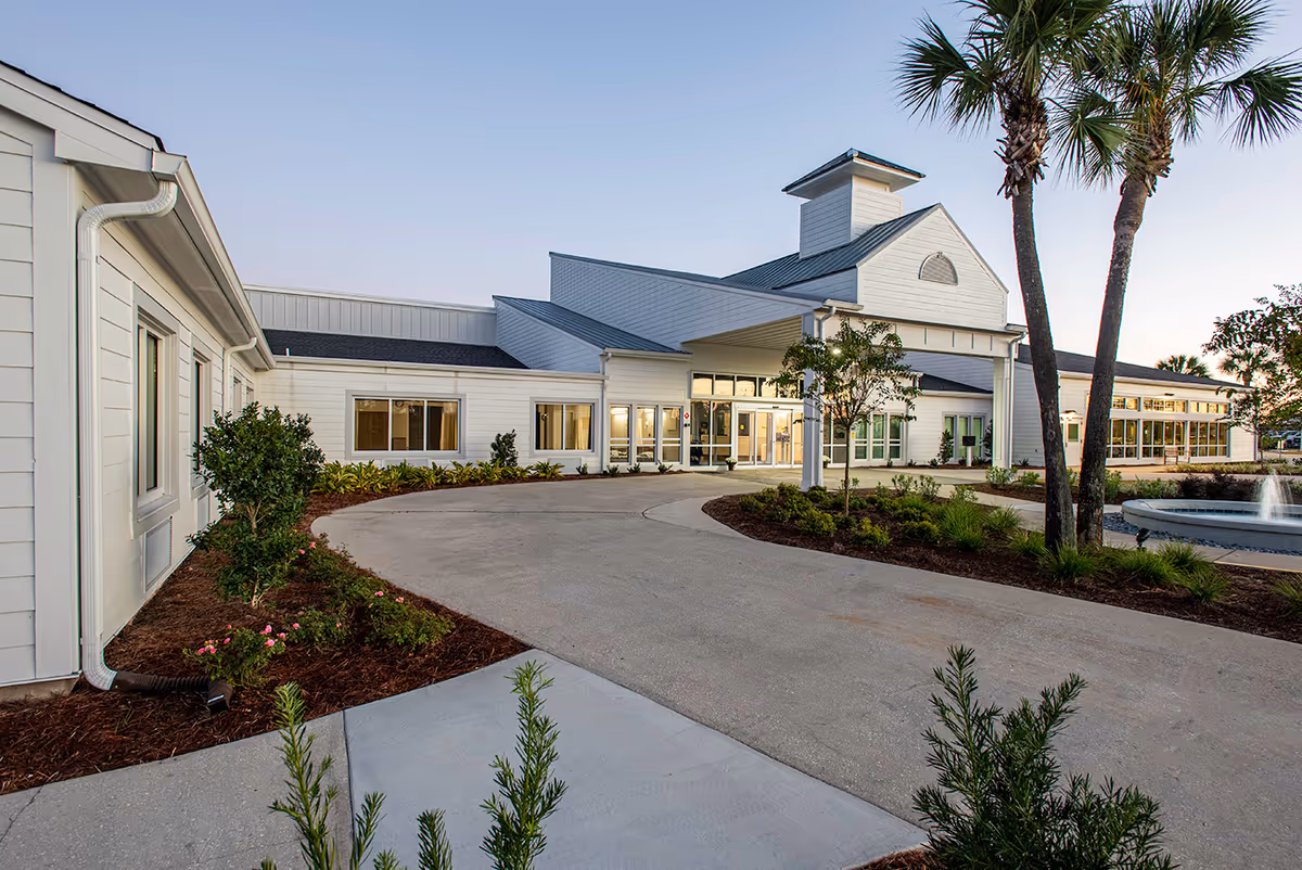 Front entrance of a white senior living facility with a circular driveway, landscaping, palm trees, and a fountain.