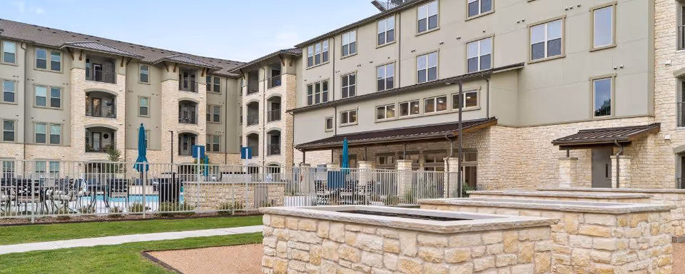 Exterior view of Preserve at Willow Park senior living facility showing a multi-story building with beige stone and stucco facade, fenced pool area with blue umbrellas, outdoor seating, and landscaped green lawn with stone planters in the foreground.