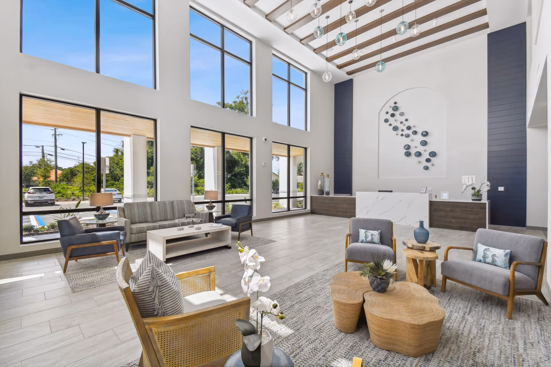 A spacious and bright senior living facility lobby with large floor-to-ceiling windows letting in natural light. The room features modern furniture including sofas, armchairs, and coffee tables arranged on area rugs. The reception desk is visible in the background with decorative wall art above it and pendant lights hanging from a high ceiling with exposed beams.
