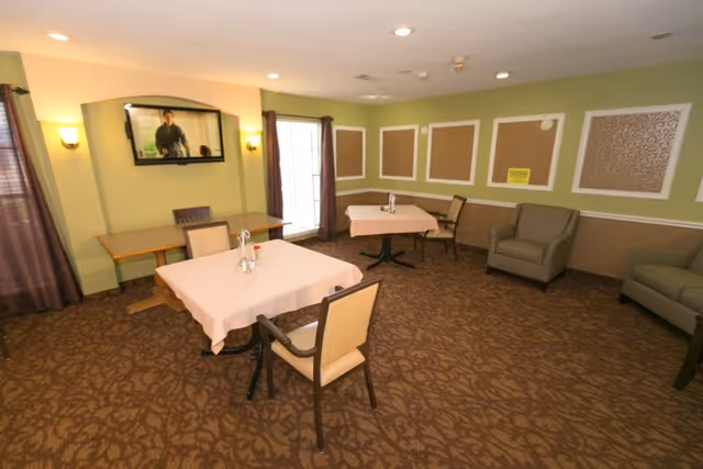 A small dining/common room with two cloth-covered tables and chairs, armchairs, a wall-mounted TV, green walls and patterned carpet.