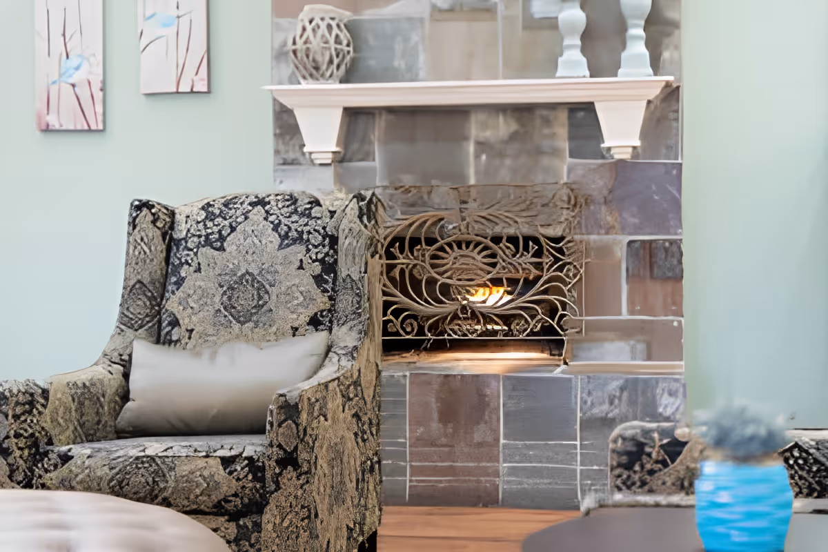 Patterned armchair with a cushion beside a lit fireplace with a decorative metal screen in a cozy living room.