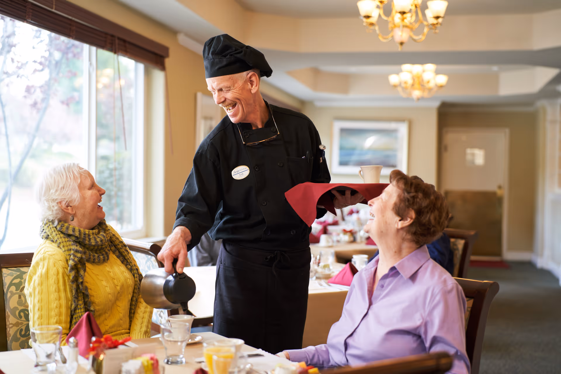 A chef in a black uniform and hat smiling and pouring coffee for two elderly women seated at a dining table in a well-lit room with large windows and chandeliers.