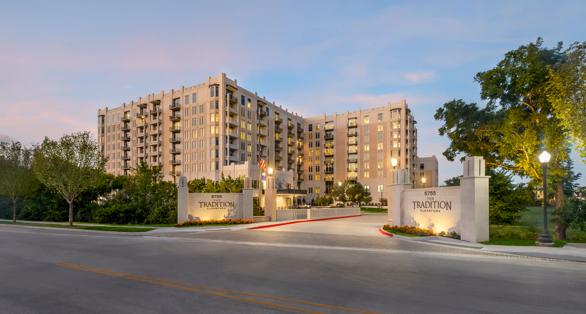Multi-story senior living building with a gated entrance, illuminated signs and landscaped grounds at dusk.
