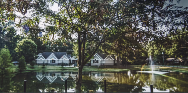 A serene outdoor scene featuring a pond with a fountain in the middle, surrounded by trees and greenery. In the background, there are several white buildings with peaked roofs reflected in the water, partially obscured by tree branches in the foreground.