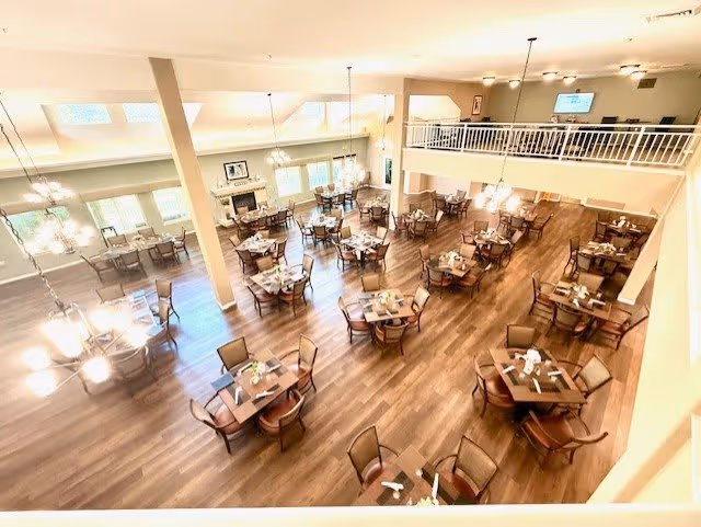 A spacious dining room with multiple square wooden tables, each surrounded by four chairs. The room has large windows allowing natural light to fill the space, wooden flooring, and modern light fixtures hanging from the ceiling. There is a mezzanine level with a railing overlooking the dining area.