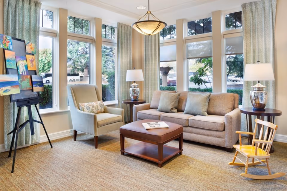 Sunlit lounge with a sofa, armchair, ottoman coffee table, easel of paintings, and large windows.