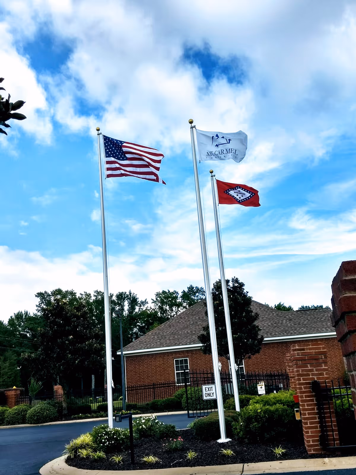 Three flagpoles with the American flag, a white Mt. Carmel flag, and a red state flag flying in front of a brick building and landscaped entrance under a partly cloudy sky.
