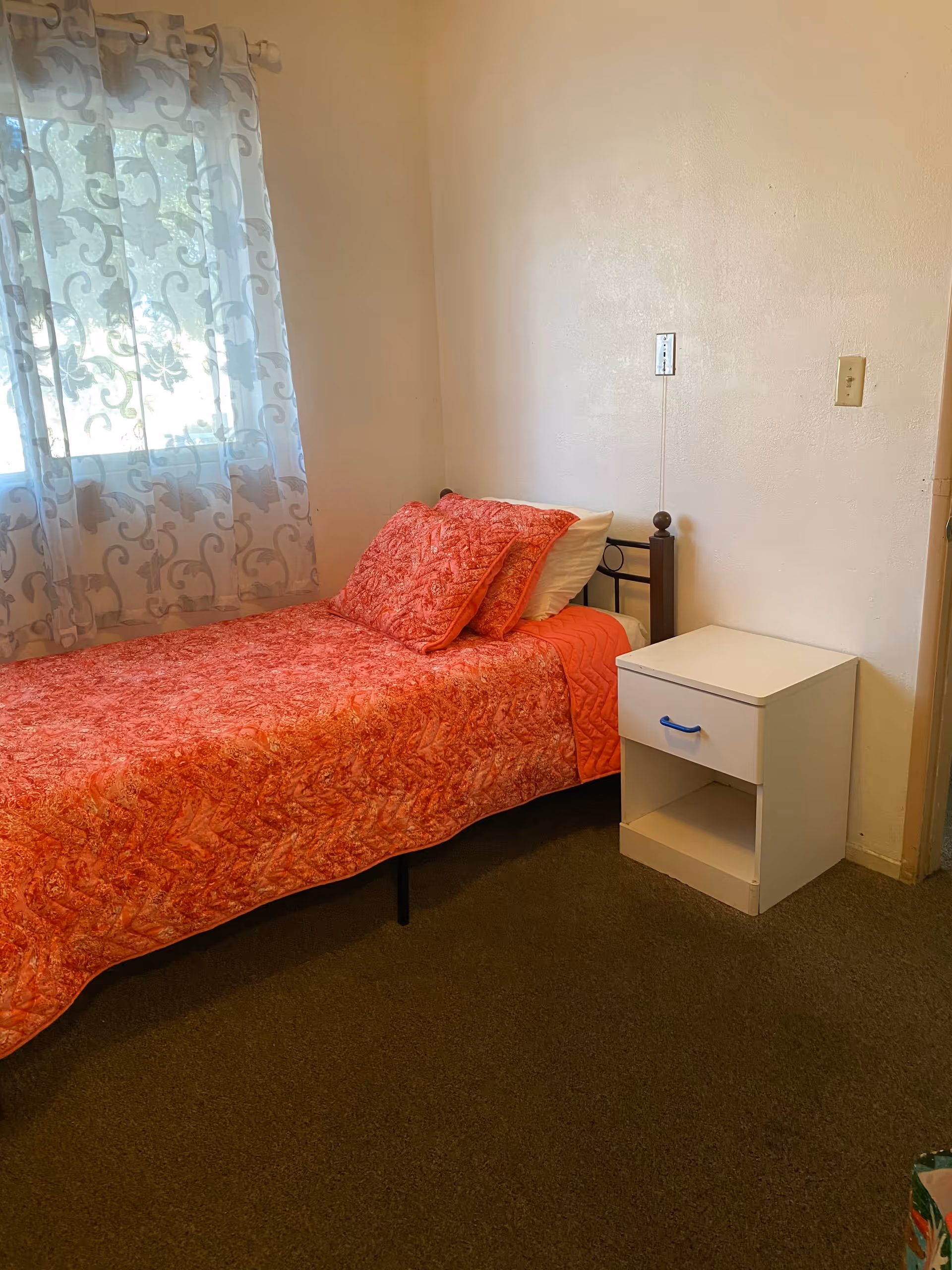 A small bedroom with a single bed covered in a red patterned quilt and matching pillows. Next to the bed is a white nightstand with a blue handle on the drawer. A window with sheer patterned curtains allows natural light into the room. The walls are plain and light-colored, and the floor is carpeted.