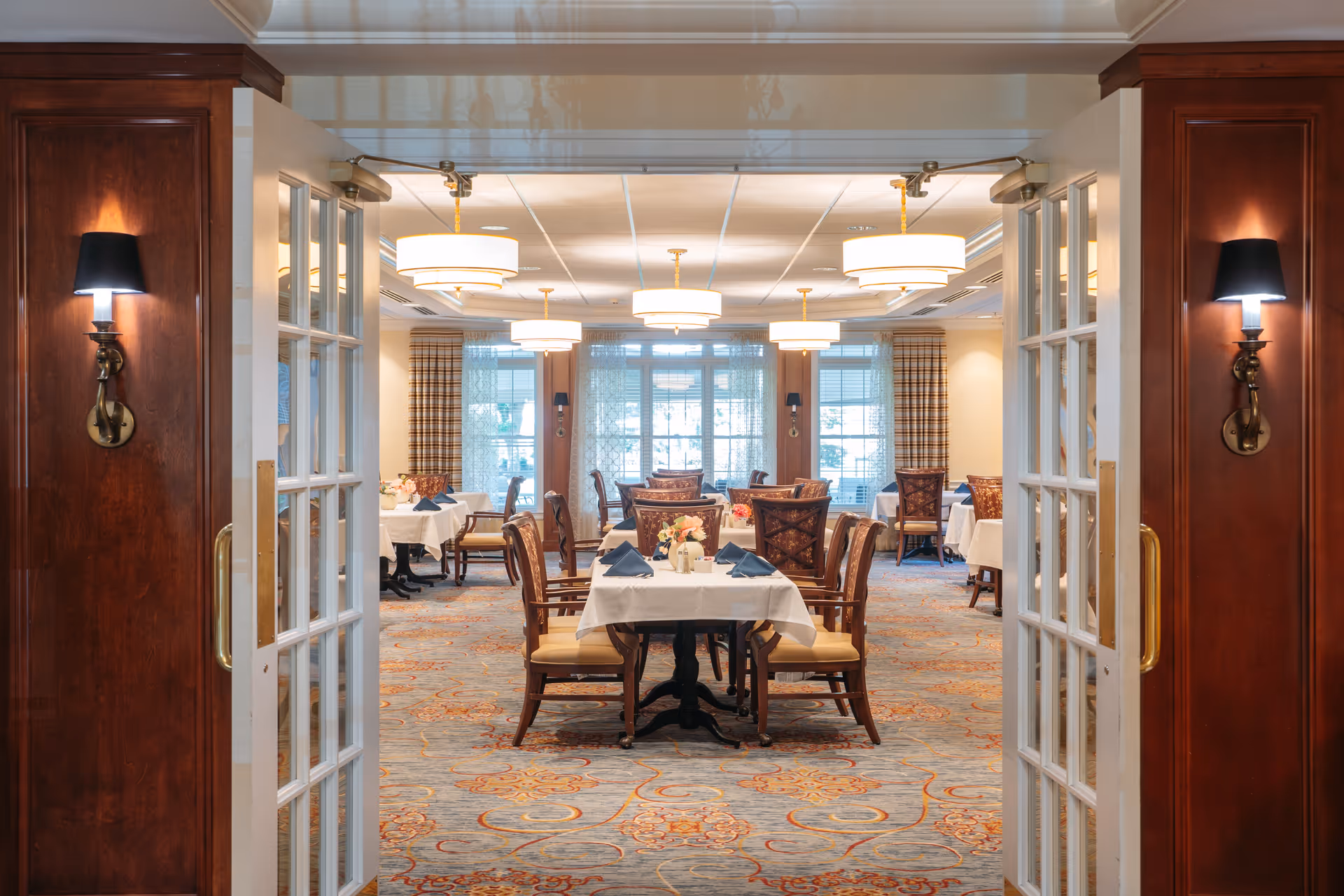 View through open double glass doors into a dining room with tables covered in white tablecloths, each set with napkins and floral centerpieces. The room features patterned carpet, wooden chairs with cushioned seats, wall sconces with black shades, and large windows with plaid curtains allowing natural light.