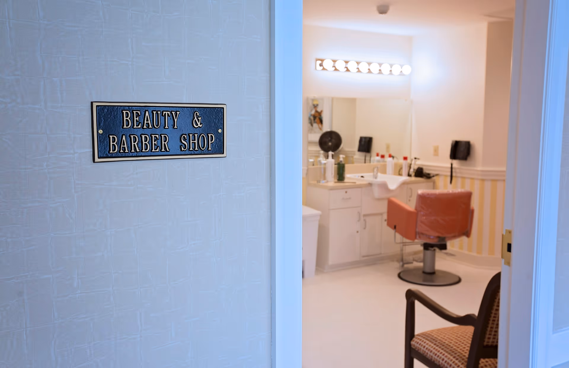 View through an open door into a beauty and barber shop room with a red barber chair, a large mirror with lights above it, and various hair care products on the counter. A sign on the wall outside the room reads 'Beauty & Barber Shop'.