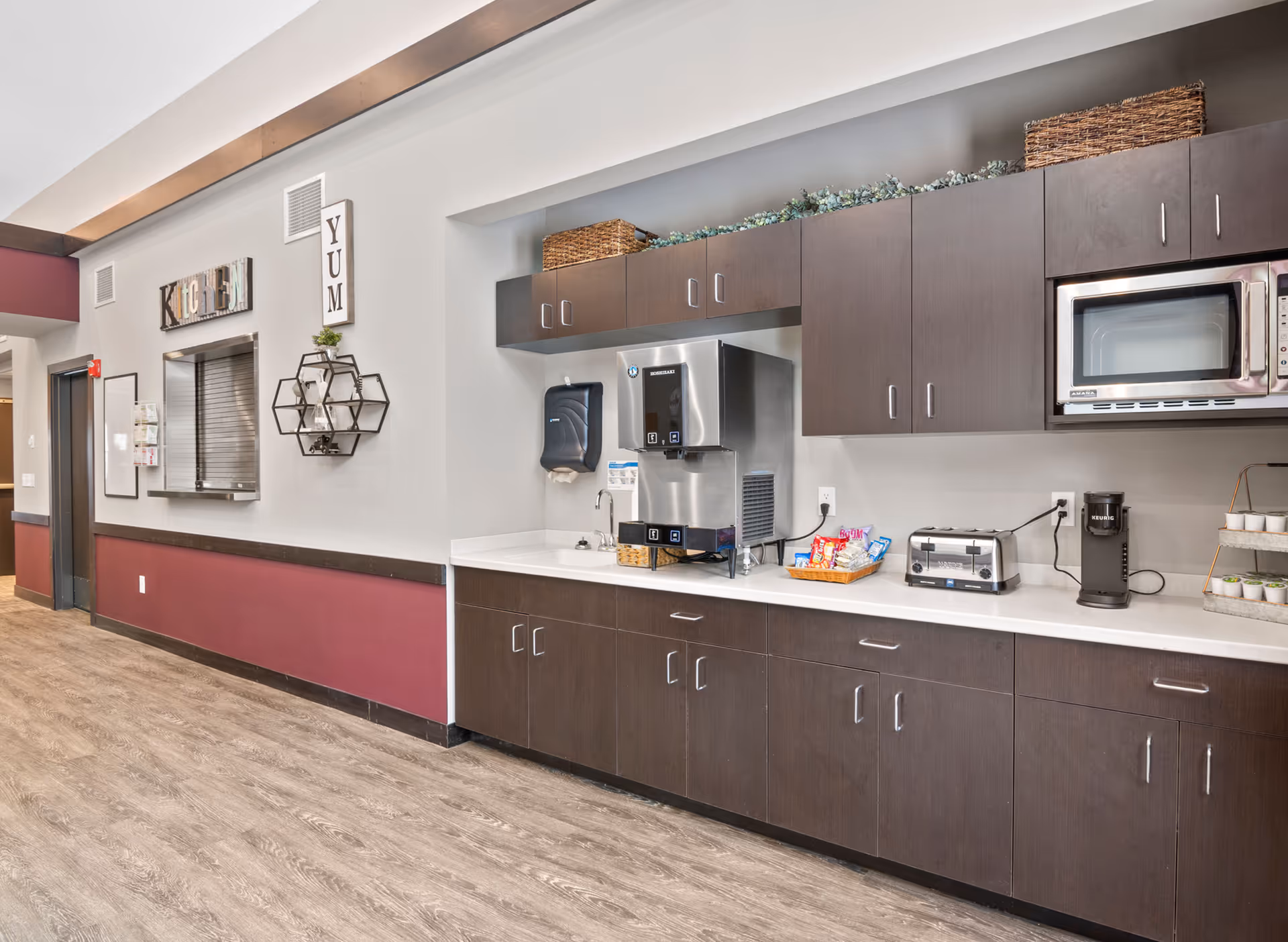 Interior view of a kitchen area in a senior living facility with dark wood cabinets, a microwave, a toaster, a coffee maker, a water dispenser, and a basket of snacks on the counter. The wall has decorative signs that say 'KITCHEN' and 'YUM'.