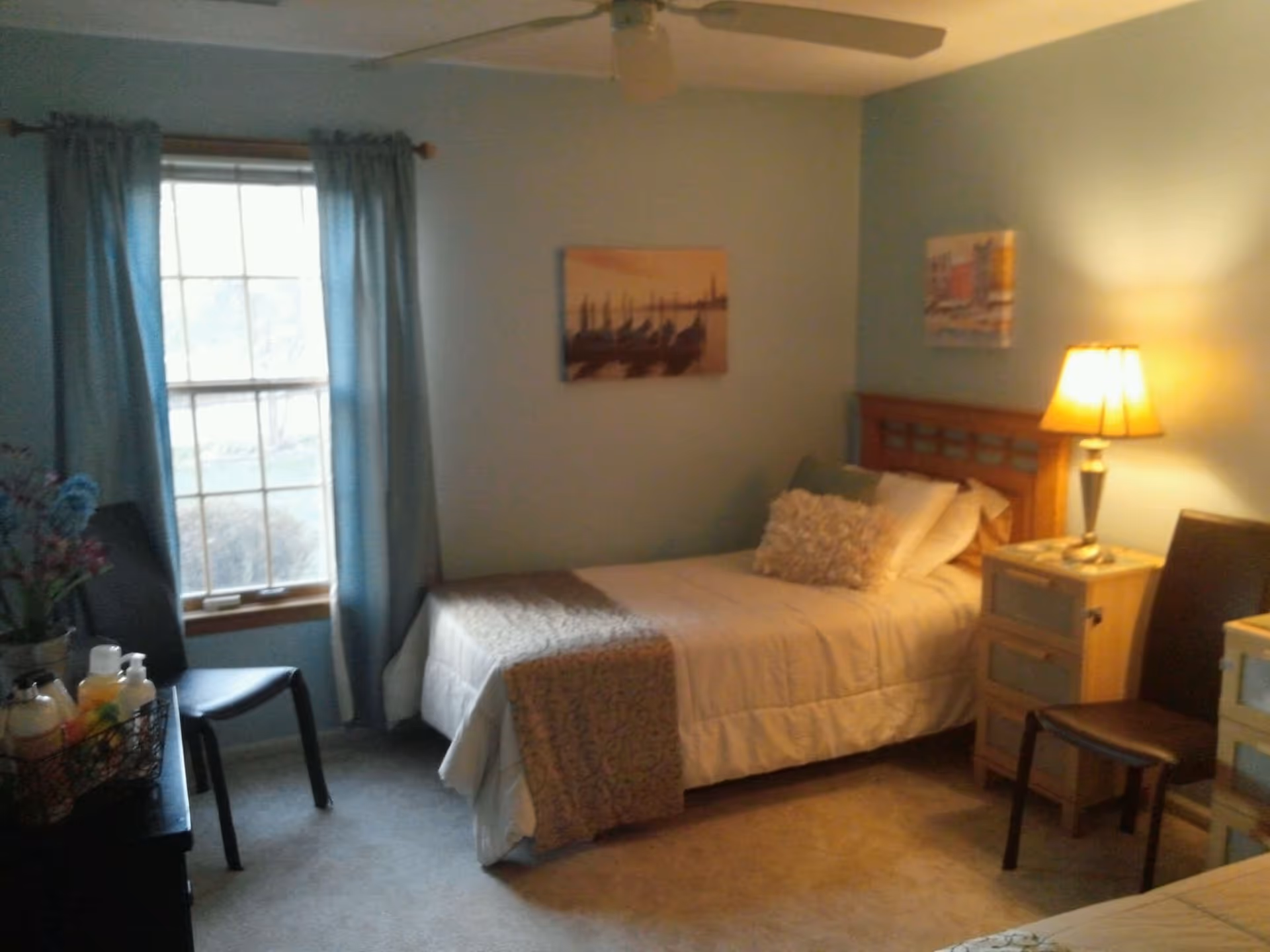 Small furnished bedroom with a single bed, nightstand and lamp, a window with blue curtains, and a chair.