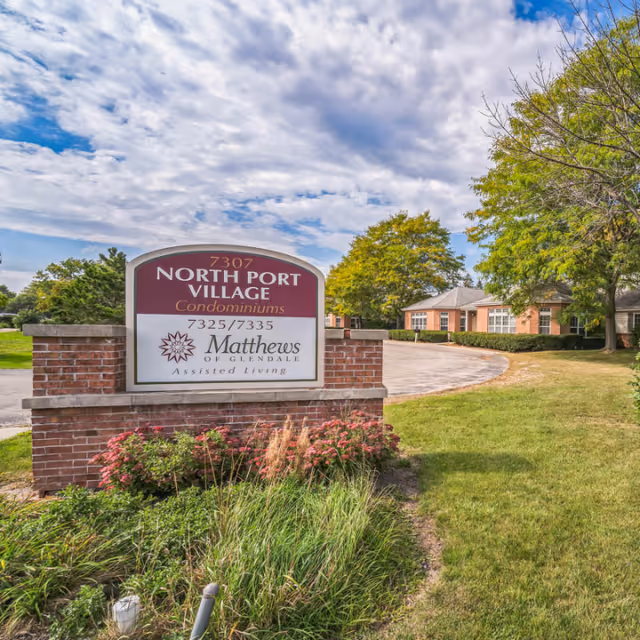 Outdoor view of a brick sign for North Port Village Condominiums and Matthews of Glendale Assisted Living, with a driveway and single-story buildings surrounded by green grass and trees under a partly cloudy sky.
