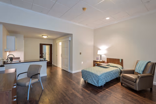 A spacious, well-lit retirement community room with a single bed covered in a blue quilt, a bedside table with a lamp, a comfortable armchair with a blue throw, a desk with a chair, and a kitchenette area with cabinets and a coffee maker. The room has wooden flooring and light-colored walls.