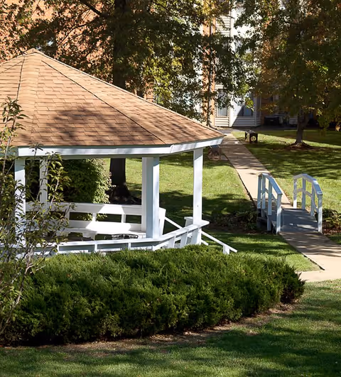 A white wooden gazebo with a brown shingled roof situated in a green grassy area with bushes and trees. A paved walkway leads to a small white wooden bridge and a building is visible in the background.