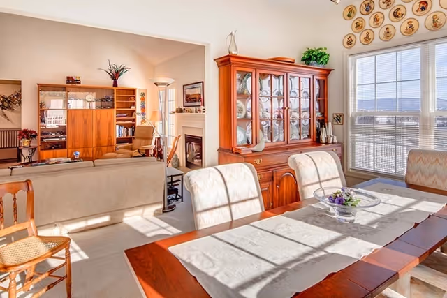 Sunlit dining room with a wooden table and upholstered chairs, a china cabinet, and an adjoining living room with sofa and fireplace.