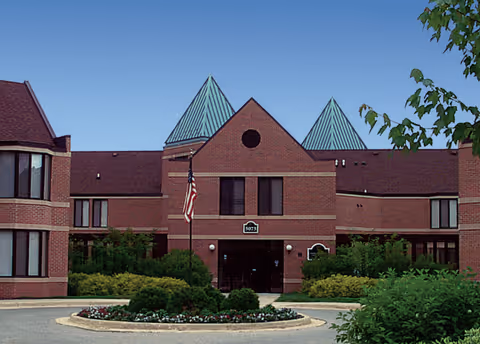 Front exterior view of Holt Heritage Village building with red brick walls, green peaked roofs, an American flag, and landscaped bushes and flowers in front.