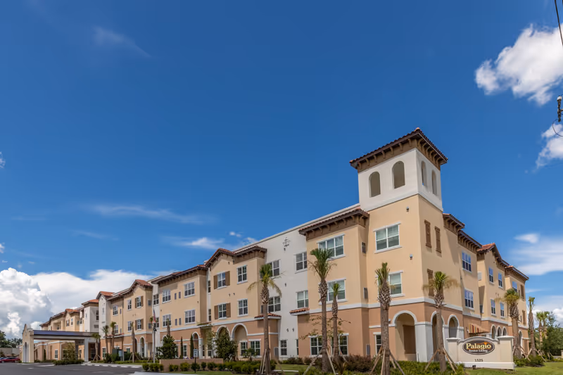 Exterior view of a large, three-story senior living facility building with a beige and light brown color scheme, palm trees planted along the front, and a clear blue sky with some clouds in the background. A sign in front reads 'Palagio Senior Living.'