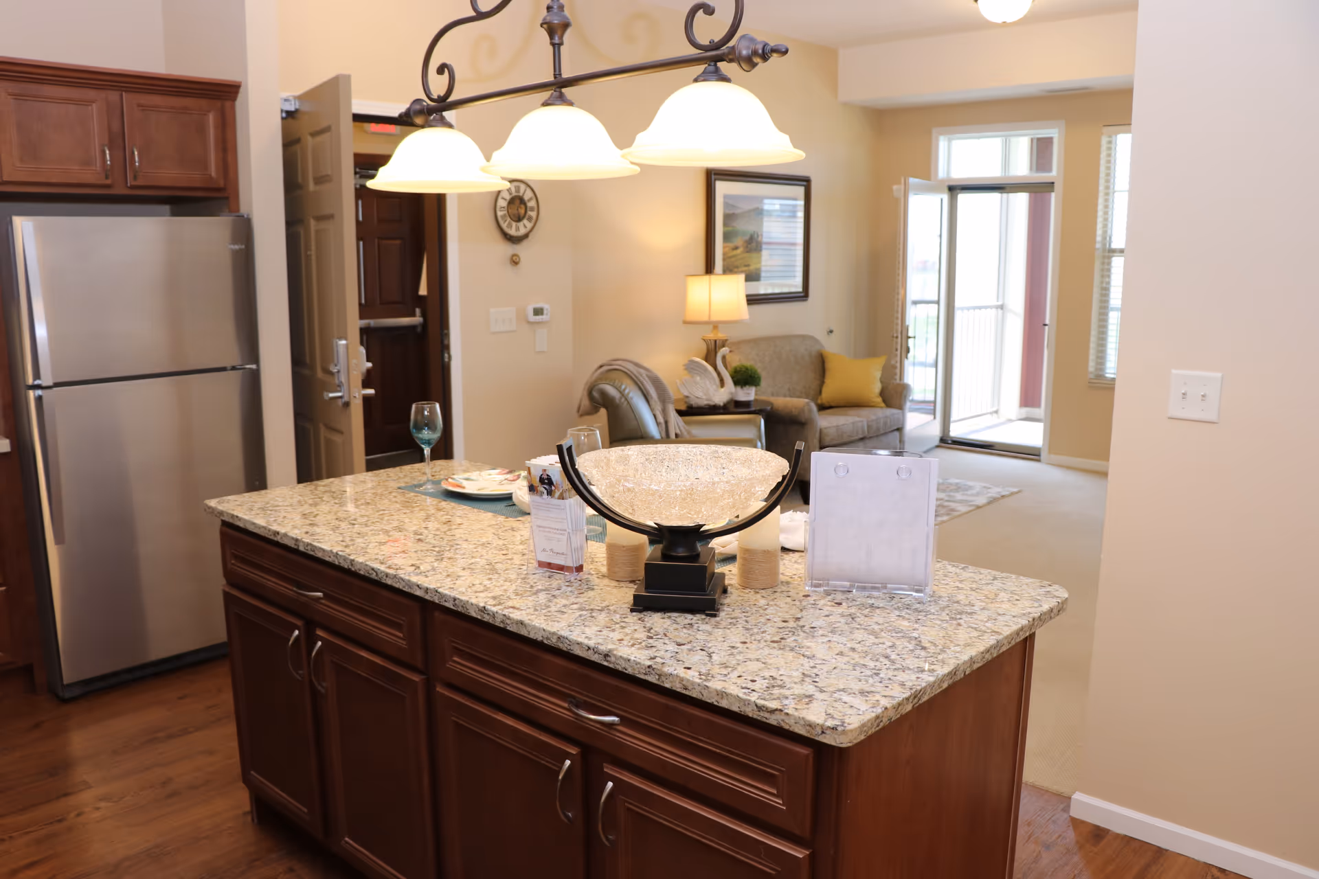 Interior view of a senior living facility showing a kitchen island with granite countertop and decorative items, a stainless steel refrigerator, wooden cabinets, and a living room area with a couch, armchair, lamp, wall clock, and a door leading to an outdoor space.