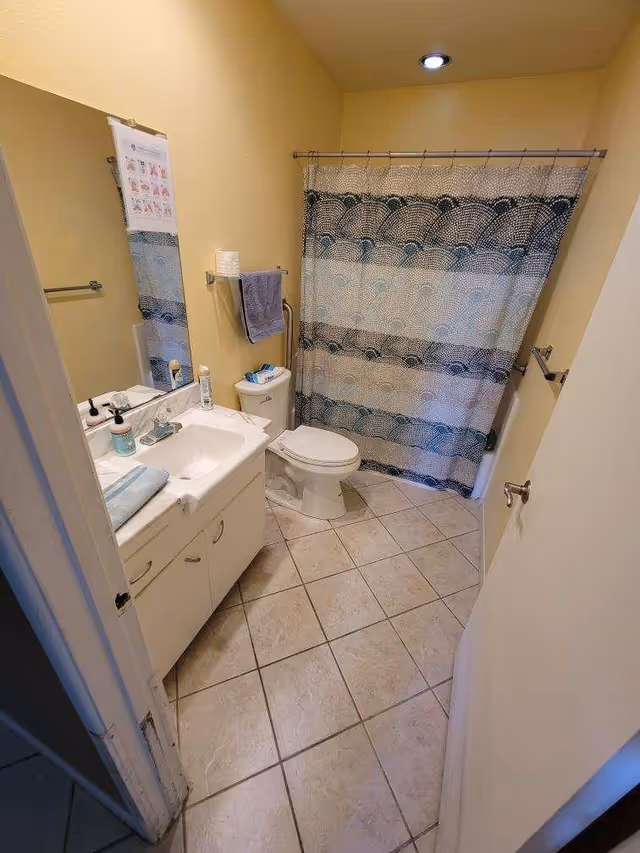 Small bathroom with a sink vanity, toilet, and a shower behind a patterned curtain on a tiled floor.
