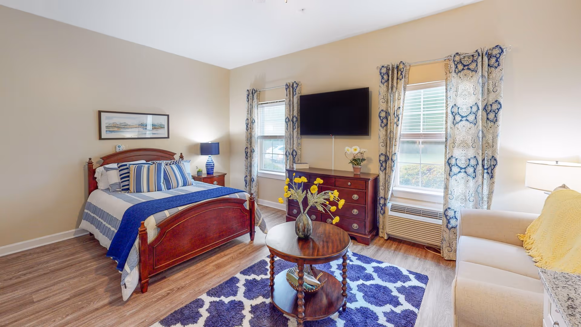 Well-lit furnished studio bedroom with a wooden bed, dresser and wall-mounted TV, sofa, round coffee table, and patterned curtains and rug.