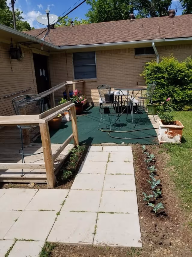 Outdoor patio area with a small table and two metal chairs on a green carpeted surface. There is a wooden ramp with handrails leading up to the patio. Several potted plants and flowers are placed around the patio. The building has a brick exterior with a window and a door. A satellite dish is mounted on the roof. The patio is surrounded by grass and a small garden bed with young plants.