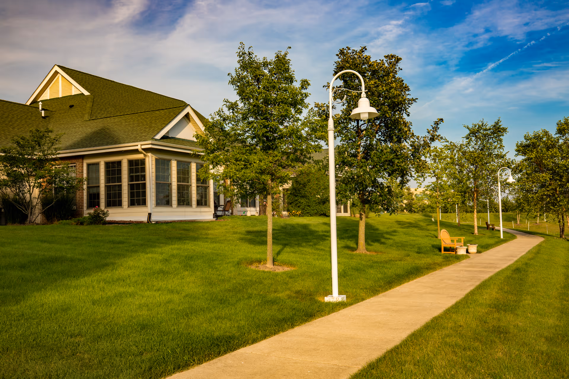 A paved walkway lined with white street lamps and benches runs alongside a green lawn with several trees. A building with large windows and a green roof is visible on the left side under a partly cloudy blue sky.