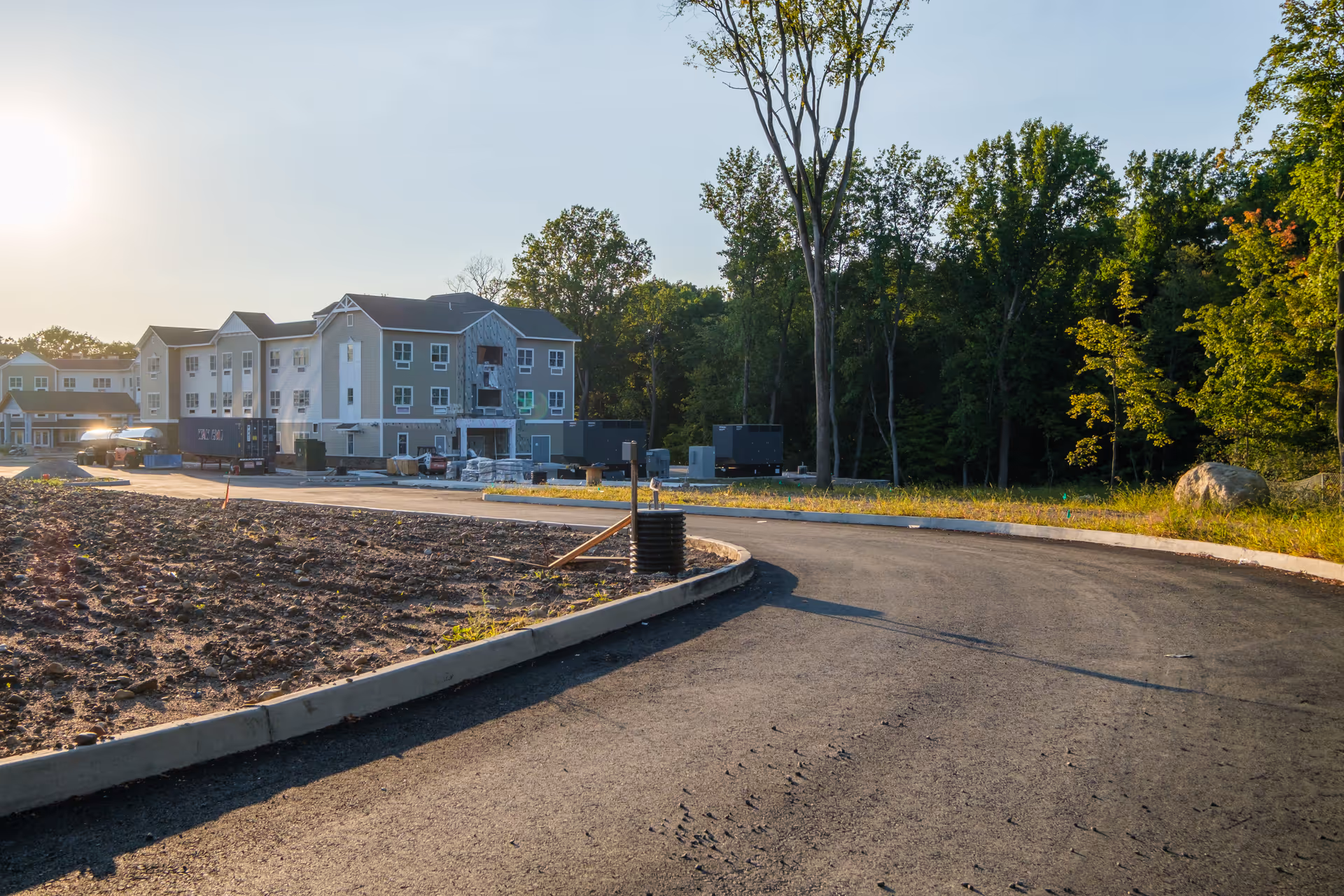 A newly paved curved road leading to a multi-story building under construction surrounded by trees and construction materials in a wooded area during daylight.