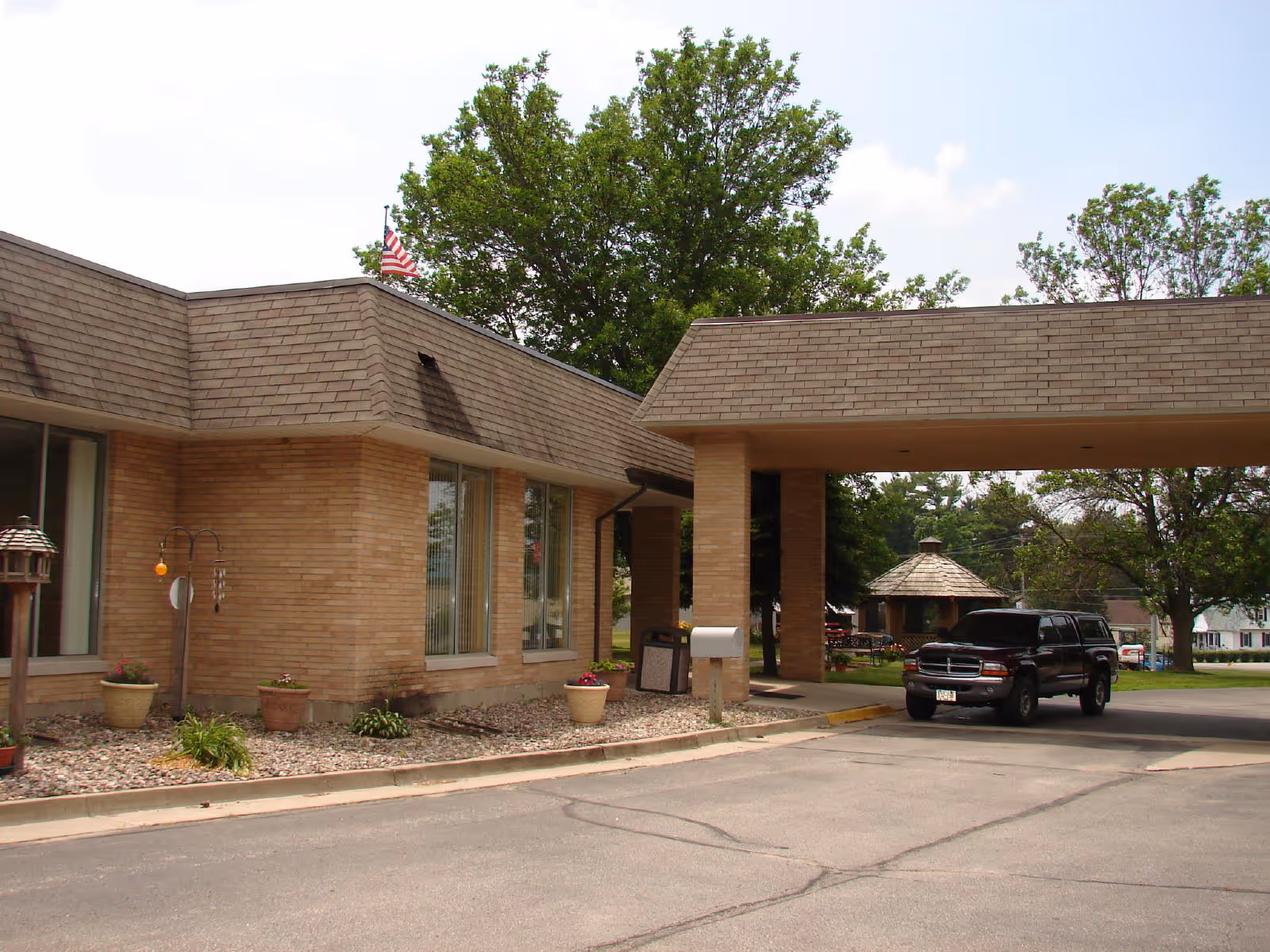 Exterior view of Marinuka Manor Nursing Home showing a brick building with large windows, a covered driveway entrance, a black SUV parked near the entrance, and trees in the background under a partly cloudy sky.