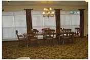 A dining room with a wooden dining table and six matching chairs on a patterned carpet. The room has large windows covered with white blinds and brown patterned curtains. A chandelier with multiple lights hangs above the table.