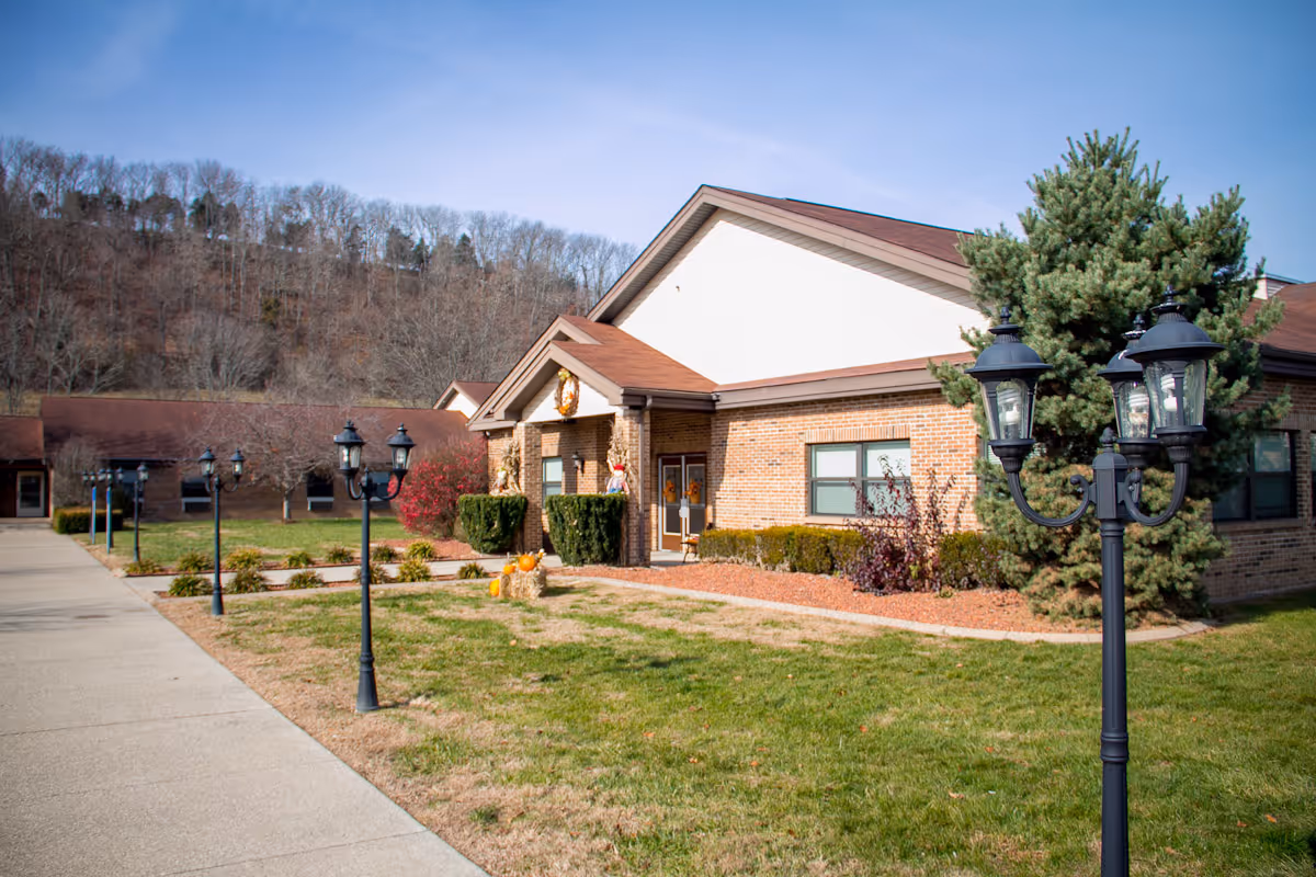 Brick one-story building front with a walkway, decorative lampposts, shrubs and a grassy lawn under a clear sky.