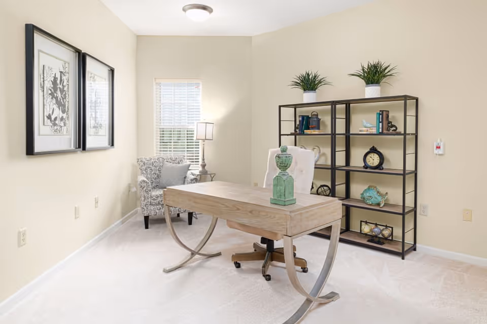A bright, neatly arranged office or study room with a wooden desk featuring curved metal legs, a cushioned office chair, a patterned armchair with a pillow, a floor lamp, two framed botanical prints on the wall, and a black metal bookshelf with decorative items and plants on top.