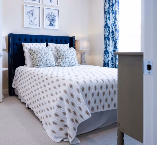 A neatly made bed with a dark blue upholstered headboard and patterned white and blue bedding in a bright bedroom. There are four framed pictures on the wall above the bed, a bedside lamp on a small table, and blue patterned curtains covering a window.