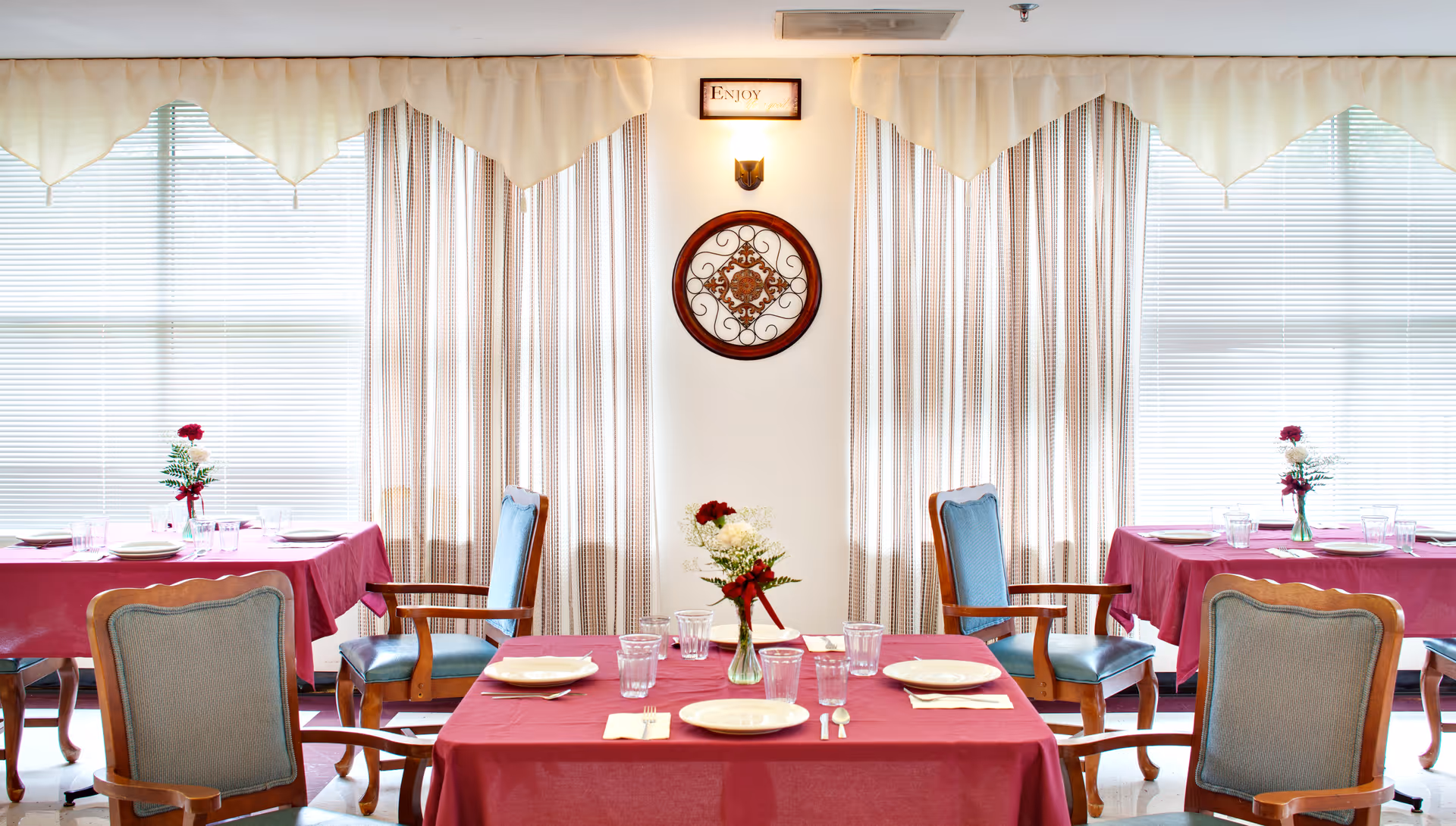 Bright dining room with tables covered in red tablecloths, place settings and floral centerpieces in front of large curtained windows.