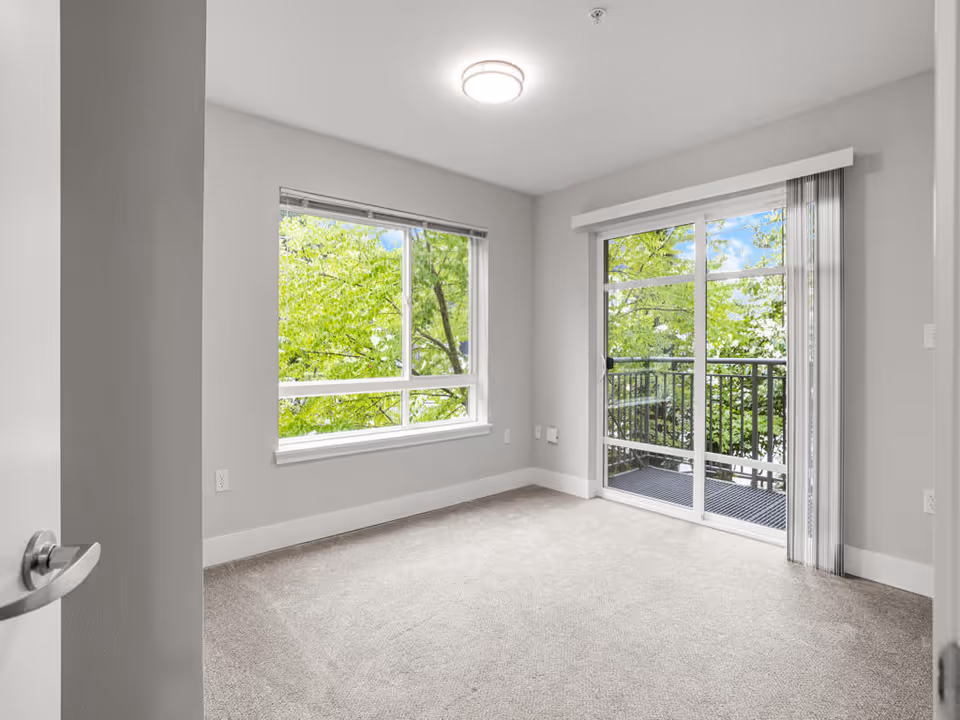 Empty room with light gray walls and carpeted floor, featuring a large window with a view of green trees on the left and a sliding glass door leading to a small balcony with black railing on the right.