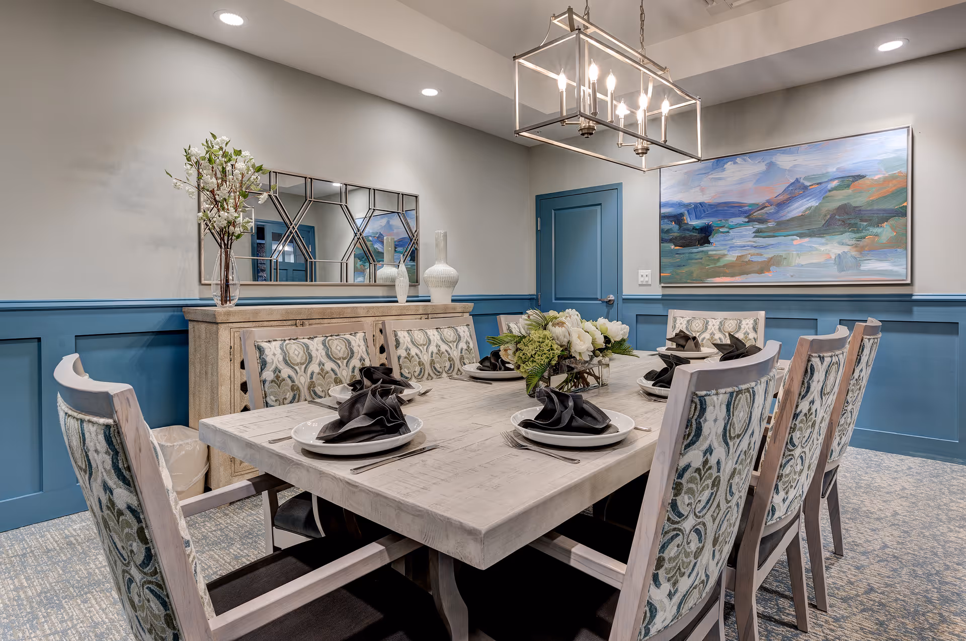 A dining room with a large rectangular wooden table set for eight people with white plates and black folded napkins. The chairs have patterned upholstery. There is a floral centerpiece on the table. The walls are painted gray with blue wainscoting and a blue door. A large abstract painting hangs on the wall, and a decorative mirror and vases are on a sideboard. A modern chandelier with multiple lights hangs above the table.