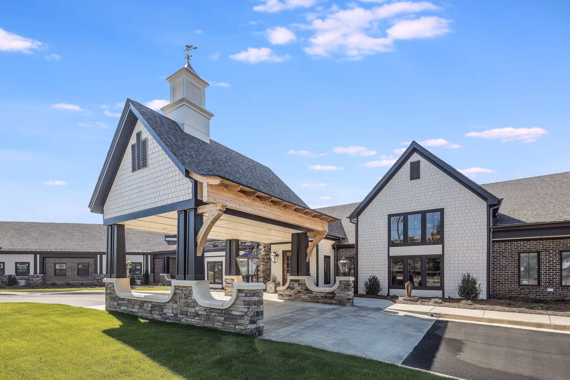 Exterior view of a senior living facility with a covered entrance supported by stone and wooden beams, adjacent to a building with large windows and a well-maintained lawn under a blue sky.