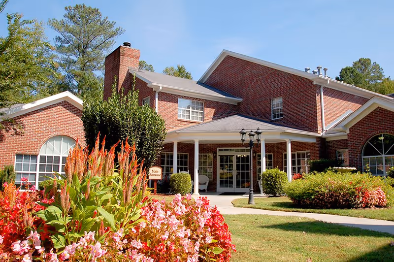Exterior view of a red brick building with white trim, featuring a covered entrance supported by white columns. The foreground has colorful flower beds with pink and red flowers, green shrubs, and a paved walkway leading to the entrance. Trees and a clear blue sky are visible in the background.