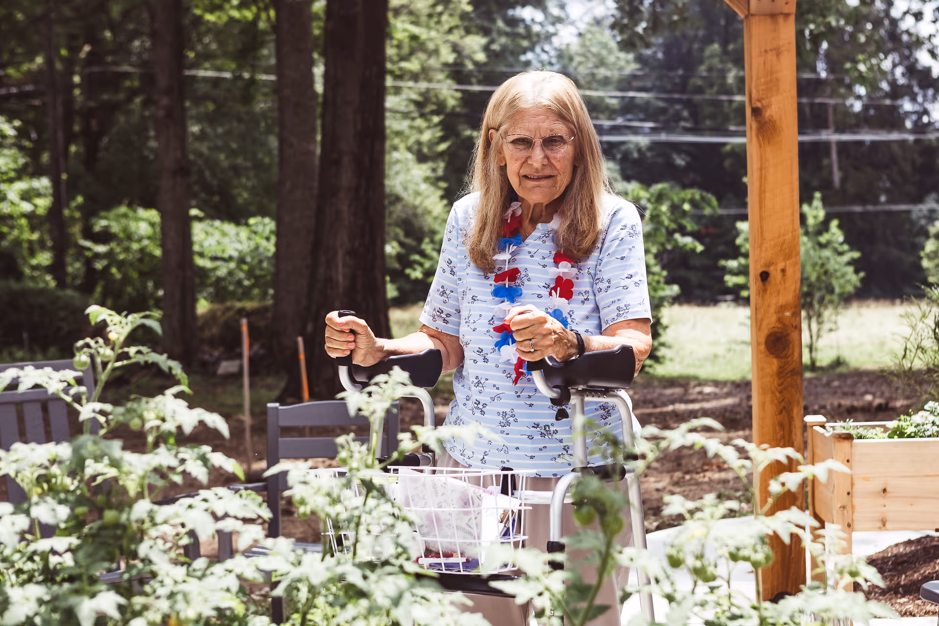 An elderly woman with long hair and glasses standing outdoors in a garden area, using a walker. She is wearing a light blue floral shirt and a red, white, and blue lei around her neck. There are green plants and trees in the background.