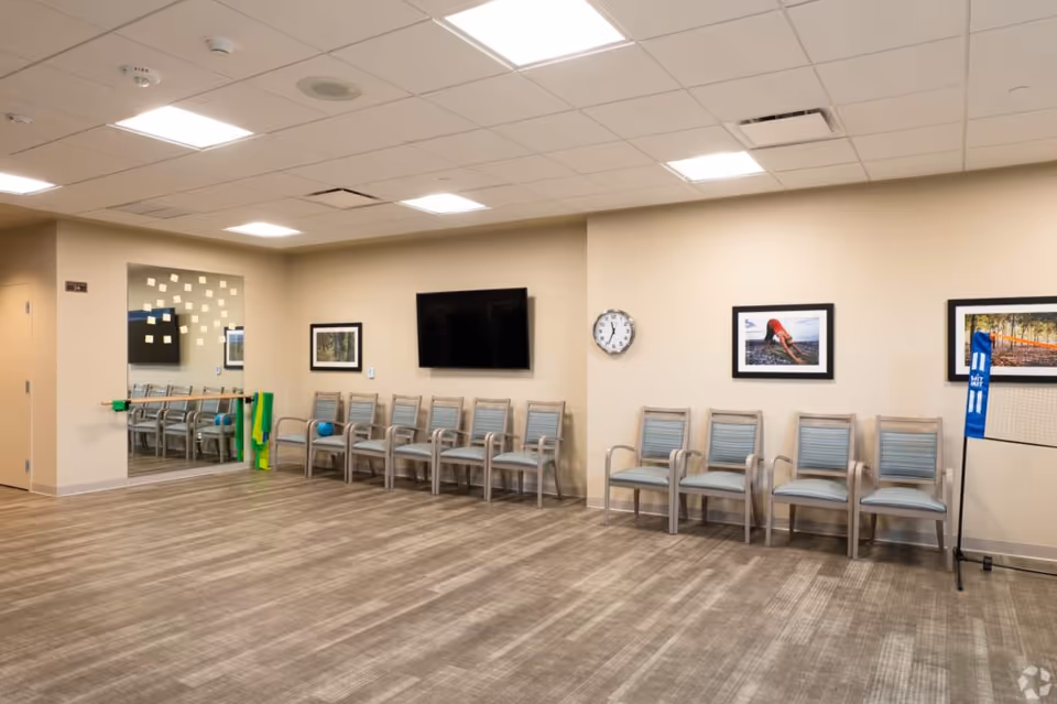 Empty multipurpose activity room with a row of chairs, a wall-mounted TV and clock, framed pictures, and a mirrored wall.