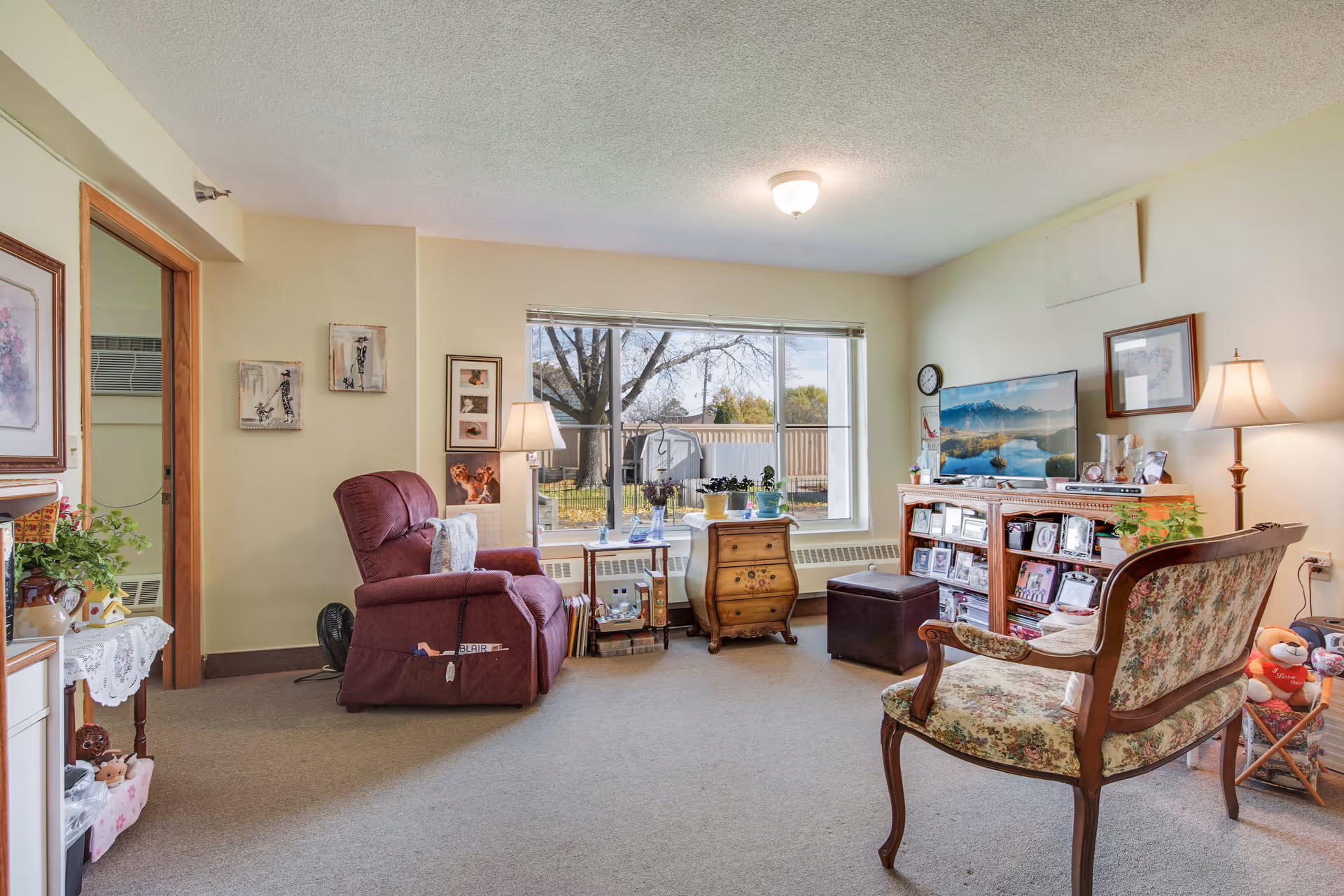 A cozy living room in an assisted living facility featuring a large window with a view of trees outside. The room contains a burgundy recliner chair, a floral upholstered wooden armchair, a wooden TV stand with a flat-screen TV, framed pictures, a small wooden chest, and two standing lamps providing warm lighting. There are various decorative items and plants placed around the room.