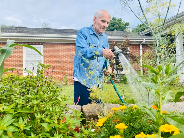 An elderly man wearing a blue denim shirt waters a garden with a hose in an outdoor area. The garden is filled with various green plants and yellow flowers. In the background, there is a brick building with white-framed windows and a cloudy sky.