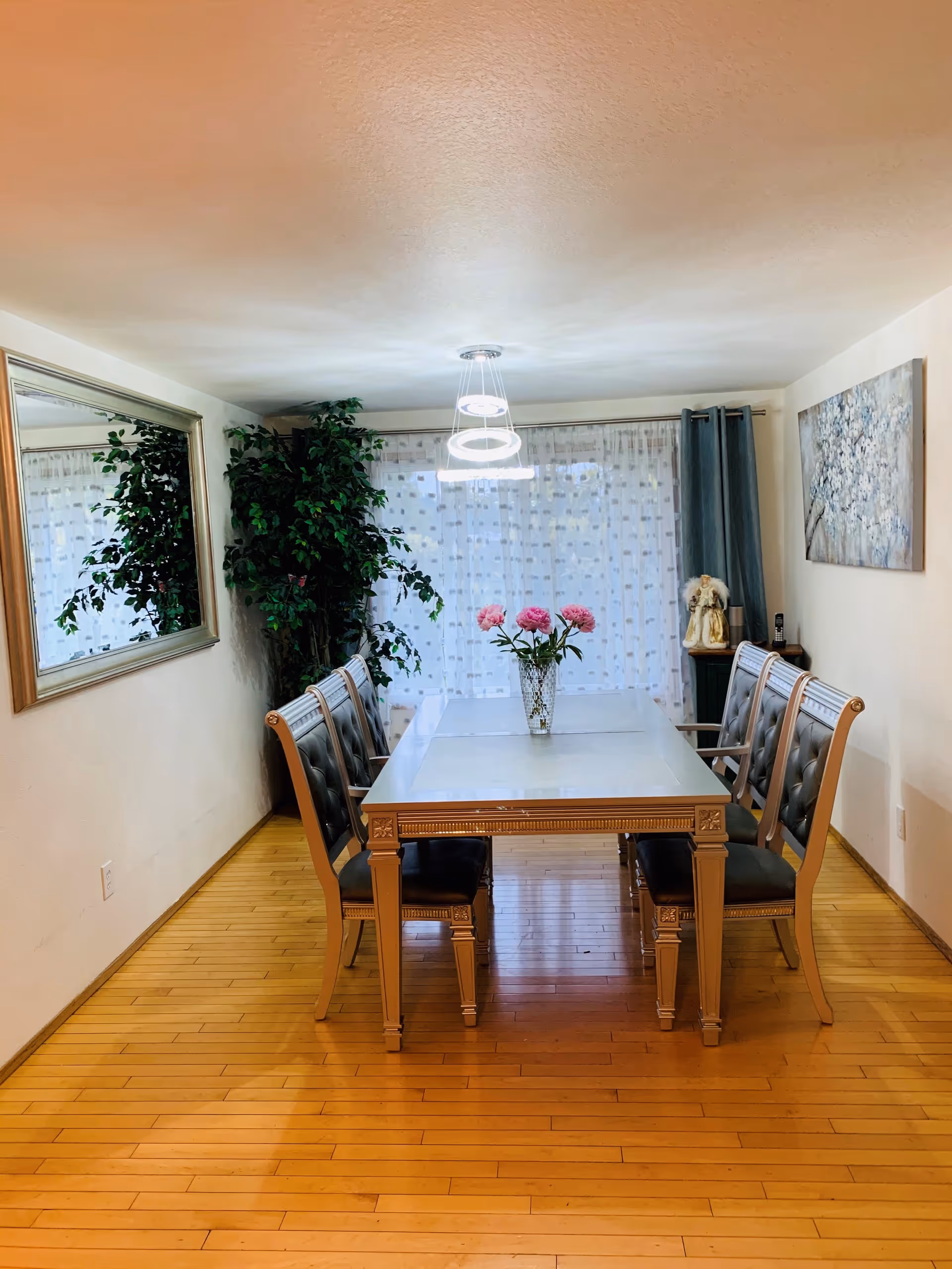 A dining room with a rectangular dining table and six chairs with dark cushioned seats and backs. A vase with pink flowers is placed in the center of the table. There is a large mirror on the left wall, a tall green plant in the corner, sheer curtains covering a large window, and a painting on the right wall. The floor is wooden and a modern circular light fixture hangs from the ceiling.