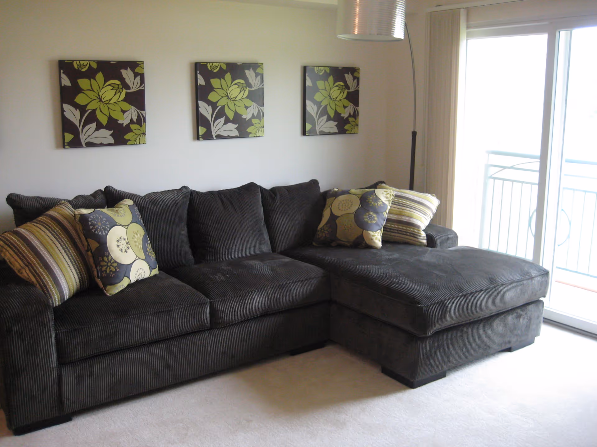 A dark gray sectional sofa with multiple decorative pillows in striped and floral patterns, positioned against a white wall with three square floral art pieces hanging above. To the right, there is a large sliding glass door leading to a balcony with a metal railing.
