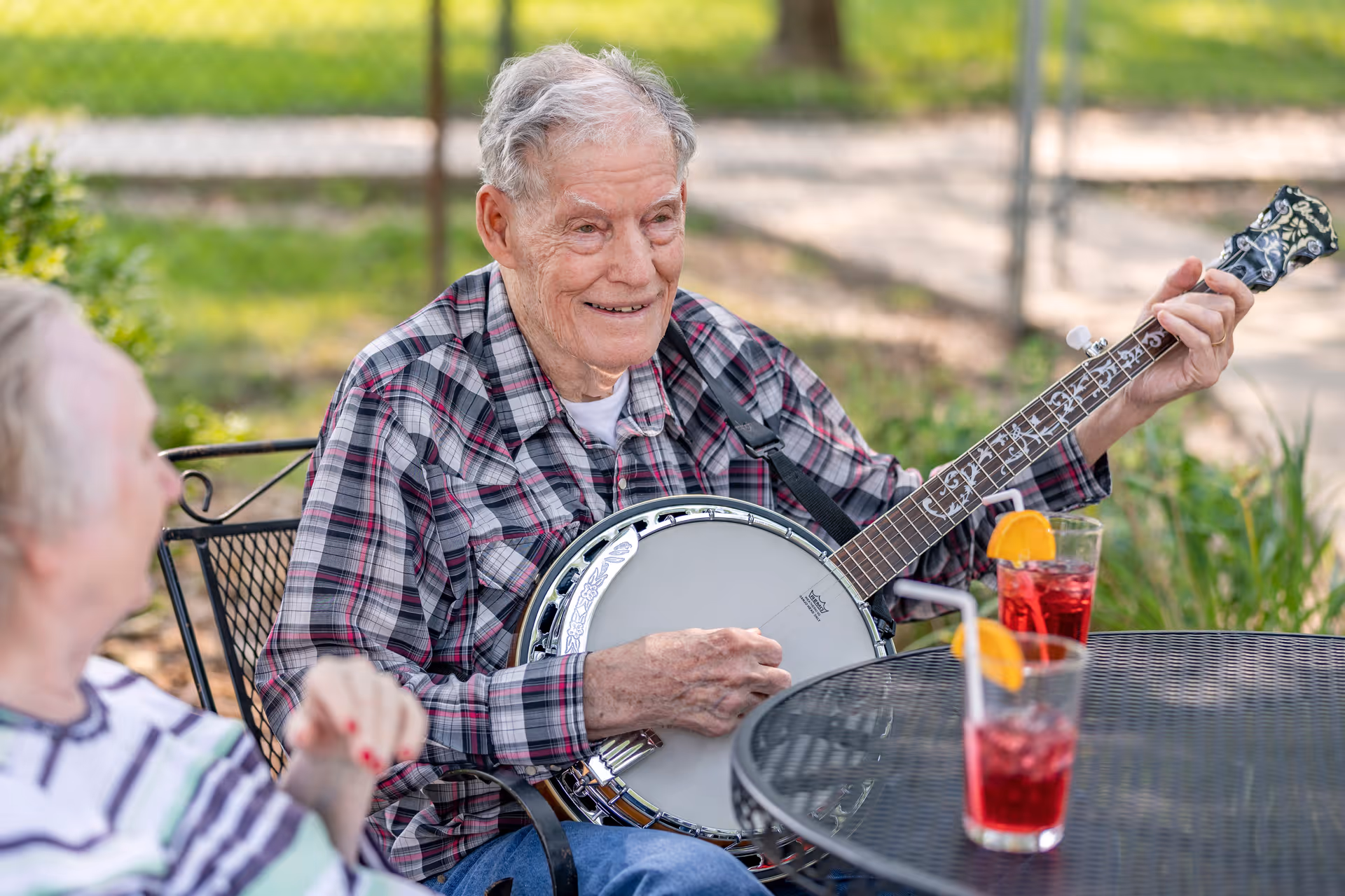An elderly man wearing a plaid shirt is sitting outdoors playing a banjo, smiling and looking at an elderly woman sitting nearby. They are seated at a black metal table with two glasses of red drink garnished with orange slices.