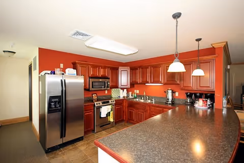 A kitchen area in Angell Park Senior Apartments featuring wooden cabinets with a reddish finish, a stainless steel refrigerator, stove, and microwave. The kitchen has a long countertop with two hanging pendant lights above it. The walls are painted a warm red color, and the floor is tiled.