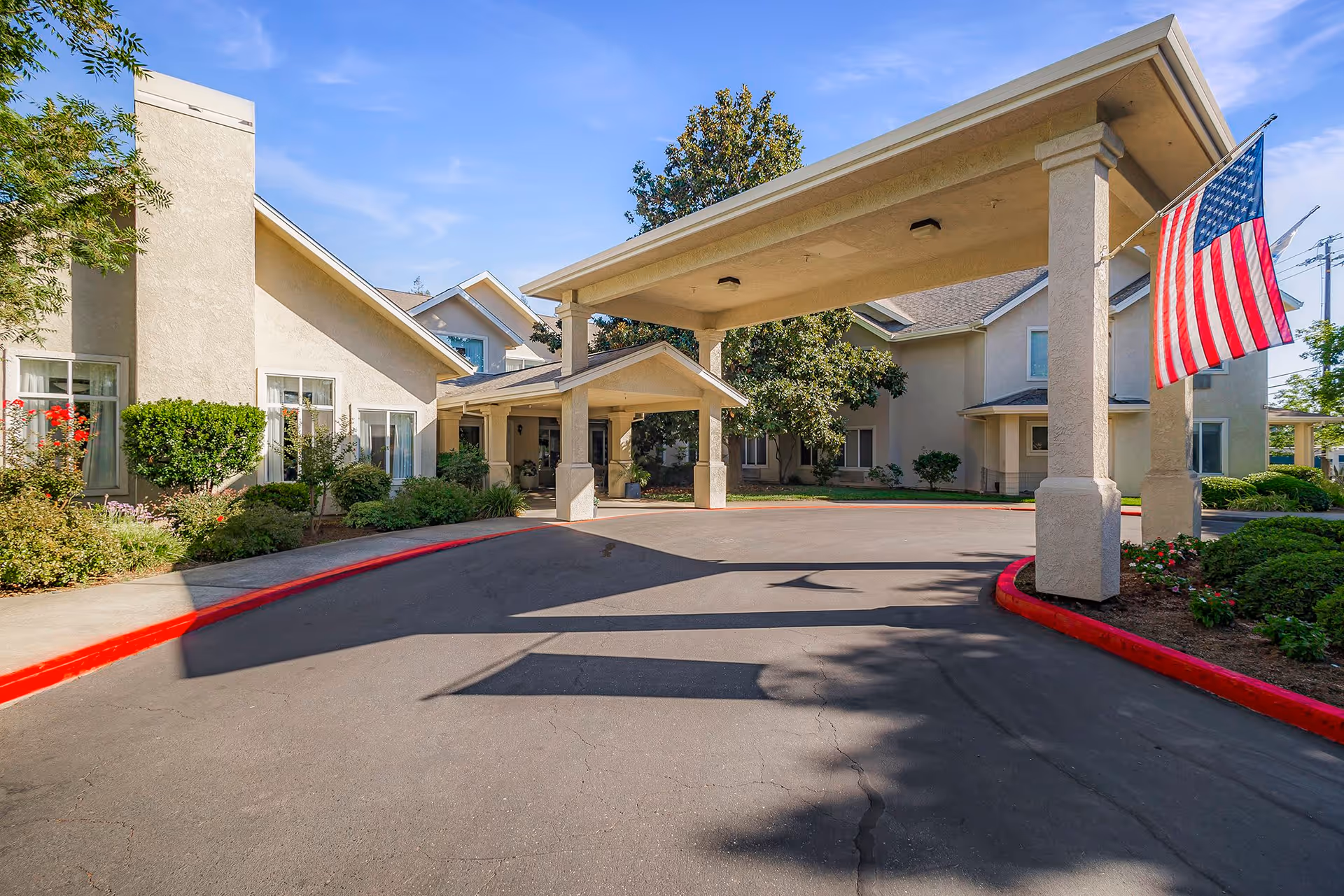 Exterior view of Marbella Chico facility entrance with a covered drop-off area supported by columns, an American flag hanging on the right side, surrounded by well-maintained landscaping and clear blue sky.